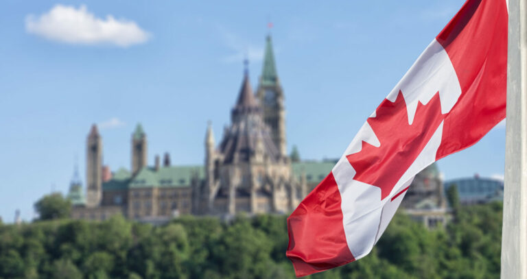 Canadian flag waving with Parliament Buildings hill and Library alternate text for this image