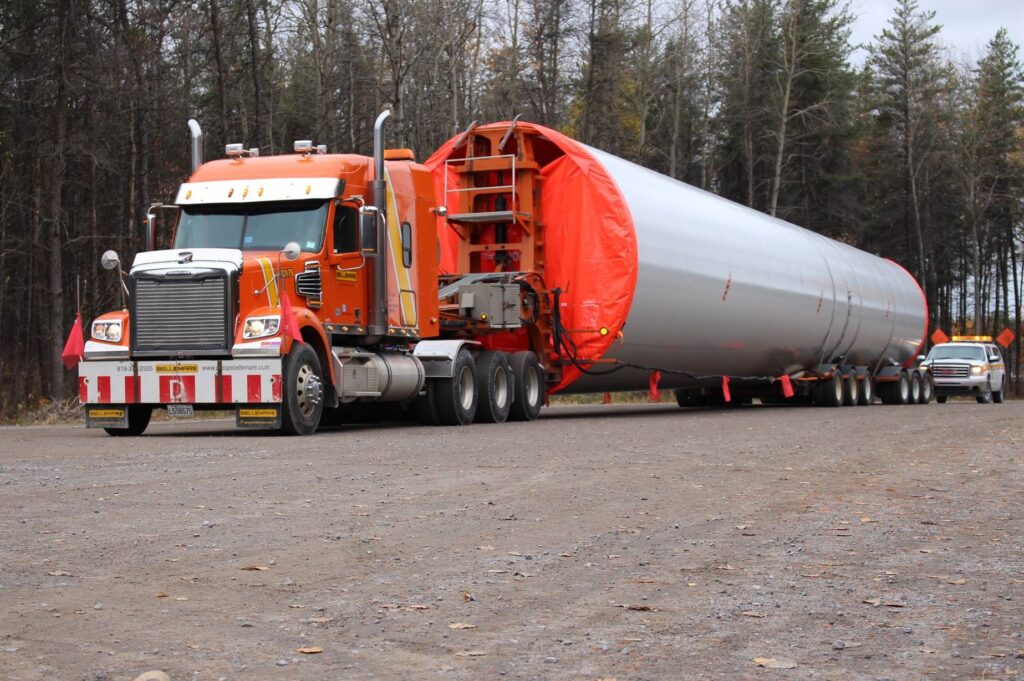 Vue de face ¾ d’un camion du Groupe Bellemare qui transporte une section d’éolienne avec véhicule d’escorte de transport hors normes à l’arrière