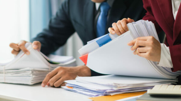 Two businesswomen are working together to search through important documents from a pile of documents on a table to verify the authenticity of the documents that were previously signed in a contract. alternate text for this image