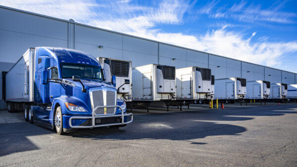 Big rig blue semi truck with refrigerator semi trailer standing in row with another semi trailers in warehouse dock gates loading cargo for the next freight alternate text for this image