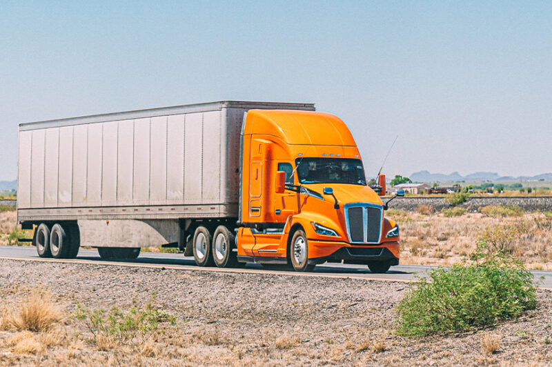Un camion orange vif tractant une longue remorque sur une route de campagne, au milieu d'un paysage aride et dégagé.