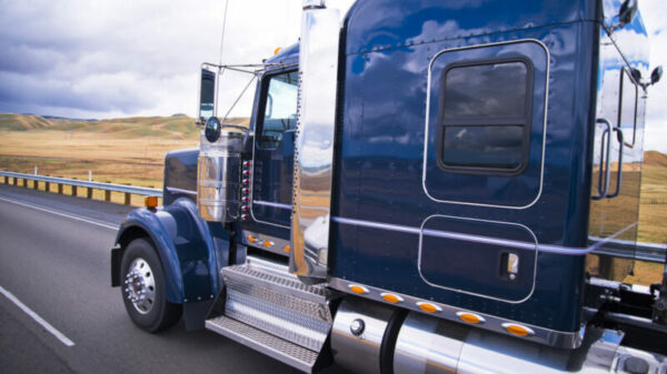 Dark blue shiny classic big rig semi truck running on the road with field on background alternate text for this image