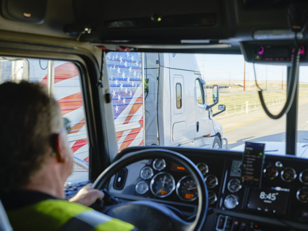 Camionneur dans sa cabine se fait dépasser par un camion dont la remorque est décorée du drapeau américain